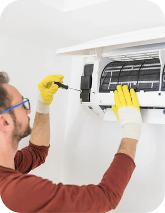 Vīrietis ar brillēm, brūnā apģērbā un dzeltenos cimdos labo gaisa kondicionieri ar skrūvgriezi, kas piestiprināts pie sienas.