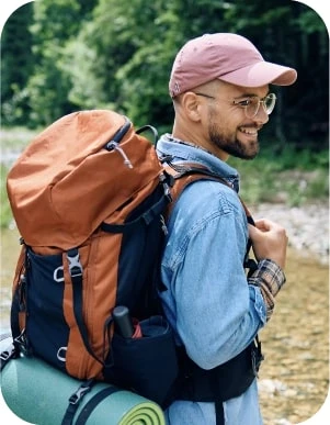 Vīrietis brillēs, rozā keponā un zilā džinsu kreklā, ar oranžu ceļojuma mugursomu un zaļu paklājiņu uz muguras, stāv pie meža malas.