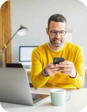 Vīrietis dzeltenā džemperī sēž pie galda ar portatīvo datoru, kafijas krūzi un mobilo telefonu rokās, fonā lapa un dators.