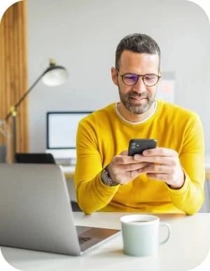 Vīrietis dzeltenā džemperī sēž pie galda ar portatīvo datoru, kafijas krūzi un mobilo telefonu rokās, fonā lapa un dators.