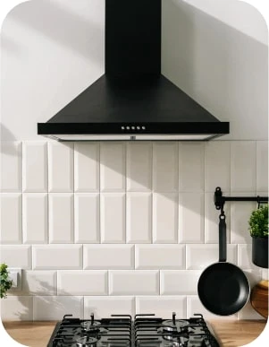 Virtuve ar melnu tvaika nosūcēju, baltiem flīzētiem sienas paneļiem, gāzes plīti un pie sienas piekarinātu pannu.