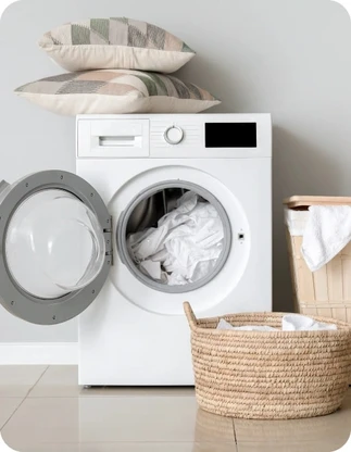 Balta veļas mašīna ar atvērtām durvīm un baltu veļu tajā, blakus divi veļas grozi ar gaišu veļu; uz veļas mašīnas virsmas novietoti divi dekoratīvi spilveni.