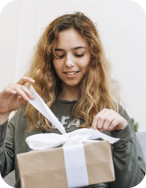 Meitene ar gariem viļņainiem matiem tumšā kreklā saiņo vaļā dāvanu kastīti, kas apsieta ar baltu lenti.