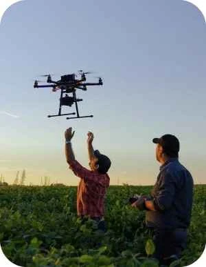 Divi vīrieši stāv laukā, viens vīrietis vada dronu, kas lido virs viņu galvām un otrs vīrietis mēģina satvert dronu.