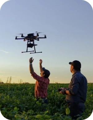 Divi vīrieši stāv laukā, viens vīrietis vada dronu, kas lido virs viņu galvām un otrs vīrietis mēģina satvert dronu.