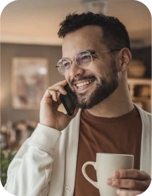 Vīrietis ar brillēm brūnā kreklā un baltā jakā runā pa mobilo telefonu, rokā turot baltu krūzi.