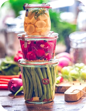 Trīs burkas ar konservētiem dārzeņiem – marinēti ķiploki ar dillēm, redīsi un zaļās pupiņas ar čili pipariem – sakrautas uz koka galda, fonā svaigi dārzeņi un virtuves piederumi.