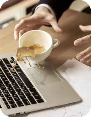 Cilvēks nejauši izlej kafiju uz portatīvā datora tastatūras, mēģinot to apturēt ar rokām; uz galda papīri ar grafikiem.