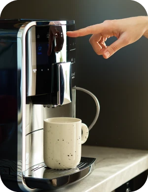 Roka spiež pogu uz kafijas automāta, zem iztekas novietota gaiša keramikas krūzīte.
