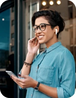 Sieviete ar tumšiem, īsiem matiem, brillēs un zilā kreklā stāv ārā, tur rokās mobilo telefonu un liek ausī baltu bezvadu austiņu.
