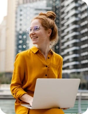 Sieviete dzeltenā kleitā sēž ārā ar portatīvo datoru klēpī; fonā augstceltnes un pilsētas ainava.