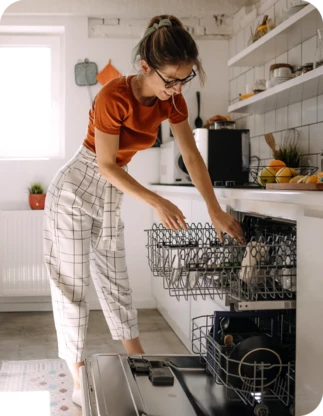 Sieviete oranžā kreklā un rūtainās biksēs, liek traukus trauku mazgājamajā mašīnā, fonā gaiša virtuve.