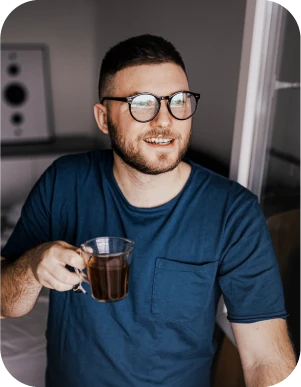 Vīrietis ar brillēm, zilā kreklā tur caurspīdīgu krūzi ar dzērienu, fonā skaļrunis un logs.