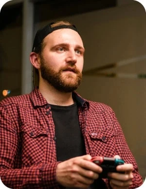 Vīrietis sarkanā, rūtainā kreklā un melnā cepurē tur rokās Nintendo Switch kontrolieri.