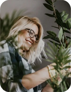 Sieviete ar gaišiem matiem, brillēm, rūtainā kreklā un dzeltenos cimdos ar ziedu rakstu kopj zaļus istabas augus, fonā telpa ar daudz augiem.