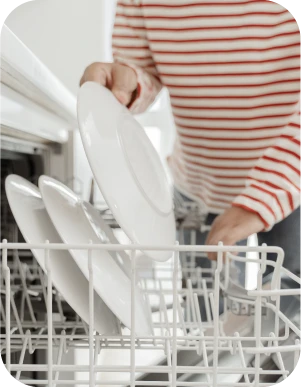 Cilvēks sarkanbalti svītrainā kreklā liek baltu šķīvi trauku mazgājamajā mašīnā.