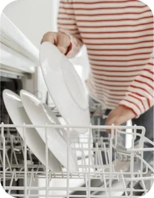 Cilvēks sarkanbalti svītrainā kreklā liek baltu šķīvi trauku mazgājamajā mašīnā.