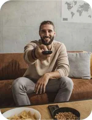 Vīrietis gaišās drēbēs sēž uz dīvāna ar televizora pulti rokā; priekšplānā galds ar čipsiem un riekstiem, fonā pie sienas pasaules karte.