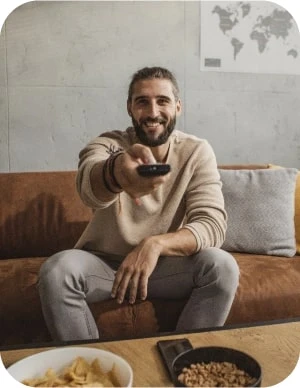 Vīrietis gaišās drēbēs sēž uz dīvāna ar televizora pulti rokā; priekšplānā galds ar čipsiem un riekstiem, fonā pie sienas pasaules karte.