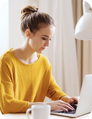 Sieviete dzeltenā džemperī sēž pie rakstāmgalda un strādā ar portatīvo datoru; blakus atrodas balta krūze un galda lampa.