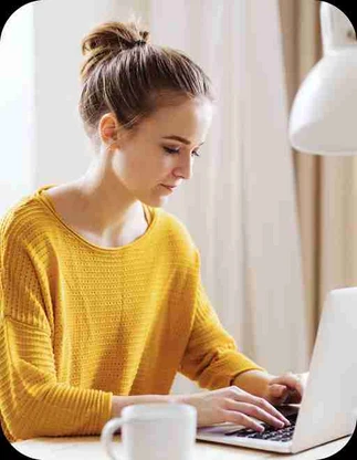 Sieviete dzeltenā džemperī sēž pie rakstāmgalda un strādā ar portatīvo datoru; blakus atrodas balta krūze un galda lampa.