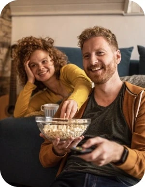 Sieviete un vīrietis sēž uz dīvāna un skatās televizoru; vīrietis tur bļodu ar popkornu, sieviete apgūlusies dīvāna atspiež galvu uz vienas rokas, bet ar otru sniedzas pēc popkorna.