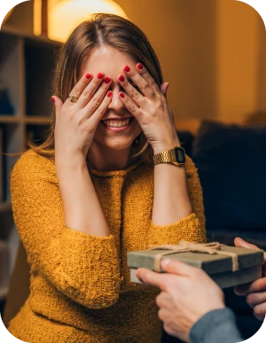 Sieviete dzeltenā džemperī piesedz acis ar rokām, kamēr kāds viņai pasniedz eleganti iesaiņotu dāvanu ar lentīti. Fonā silta un mājīga telpa.