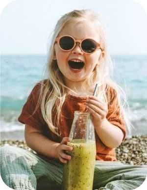 Maza meitene ar gariem gaišiem matiem un saulesbrillēm uz acīm sēž pludmalē un dzer zaļu dzērienu no stikla pudeles caur salmiņu.