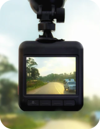 Automašīnas videoreģistrators ar ekrānu, kurā redzams dienas skats uz divvirzienu ceļu ar priekšā braucošu auto un kokiem ceļa malās.