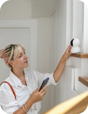 Sieviete baltā kreklā tur rokās mobilo telefonu un pieskaras nelielai video novērošanas kamerai, kas novietota uz plaukta pie sienas, fons gaišs.