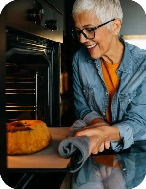 Sieviete ar brillēm, zilā džinsa kreklā, stāv pie atvērtas cepeškrāsns un ņem ārā svaigi ceptu kēksu, turot cepeškrāsns pannu ar dvieli.