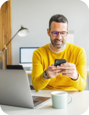 Vīrietis dzeltenā džemperī sēž pie galda ar portatīvo datoru, kafijas krūzi un mobilo telefonu rokās, fonā lapa un dators.