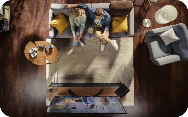 Skats no augšas uz dzīvojamo istabu, kur divi cilvēki sēž uz pelēka dīvāna ar dzelteniem un brūnu spilvenu, skatoties televizoru; pie dīvāna ir apaļš koka kafijas galdiņš, stāvlampa, pelēks krēsls ar baltu spilvenu, grīdu sedz bēšs paklājs.