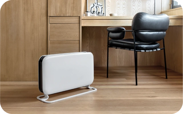 Uz koka grīdas novietots balts, taisnstūrveida sildītājs ar noapaļotiem stūriem un metāla statīvu; fonā brūns skapis, rakstāmgalds un melns ādas krēsls ar metāla kājām.