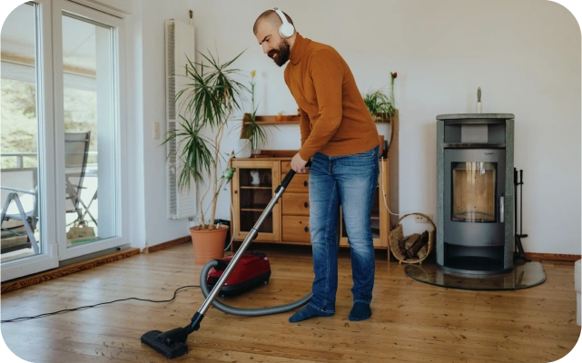 Vīrietis brūnā džemperī un džinsos tīra viesistabas koka grīdu ar putekļu sūcēju; kreisajā pusē liels logs, fonā telpaugs, koka plaukts un krāsns ar stikla durvīm.