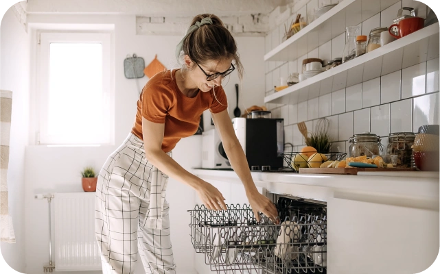 Sieviete oranžā kreklā un rūtainās biksēs, liek traukus trauku mazgājamajā mašīnā, fonā gaiša virtuve.