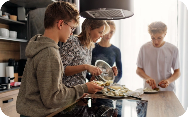 Četri cilvēki virtuvē rosās ap virtuves salu — viens tur šķīvi ar ēdienu, cits tur katla vāku, trešais darbojas ar ēdienu uz šķīvja; fonā plīts, tvaika nosūcējs un plaukti ar traukiem.