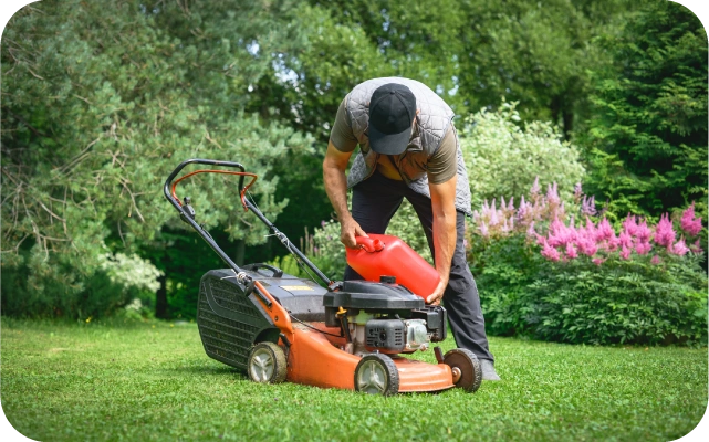 Vīrietis pieliecies lej degvielu no sarkanas kanniņas oranžas un melnas krāsas zāles pļāvējā; fonā zaļš zāliens, koki un puķu dobes.