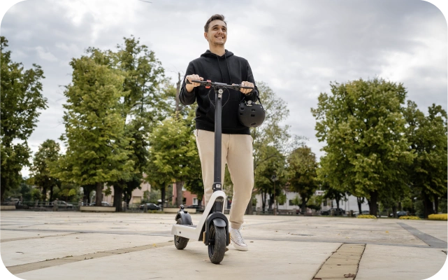 Vīrietis stāv uz elektriskā skrejriteņa pilsētas parkā vai pilsētvidē, turot stūri rokās un pakarinātu aizsargķiveri uz vienas rokas; fonā koki, bruģēta virsma un ēku fragmenti.