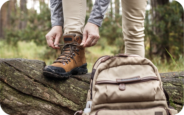 Cilvēks uzlicis kāju uz nokrituša koka stumbra mežā, sašņorē brūnus pārgājienu zābakus; priekšplānā bēša mugursoma, fonā koki un krūmi.