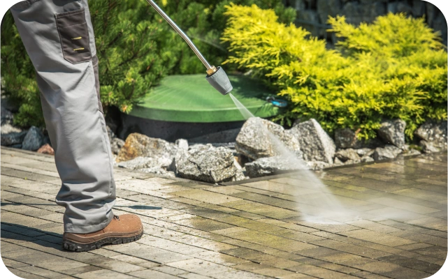 Cilvēks pelēkās biksēs un brūnos apavos ar augstspiediena mazgātāju tīra bruģētu virsmu dārzā, fonā zaļi augi un krūms.