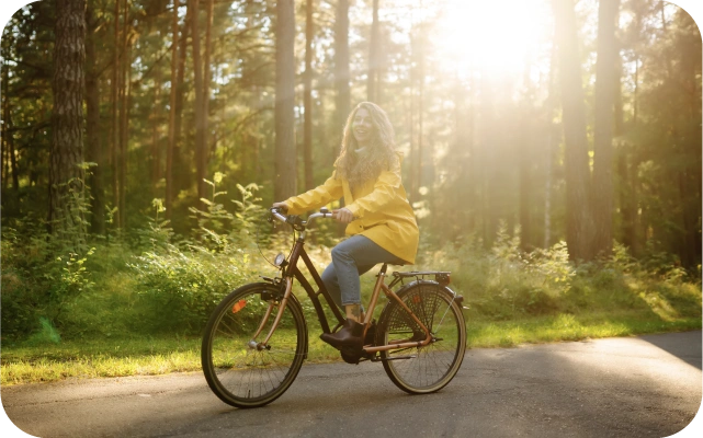 Sieviete dzeltenā mētelī brauc ar velosipēdu pa asfaltētu ceļu caur mežu, kur saules stari spraucas cauri koku lapotnei.
