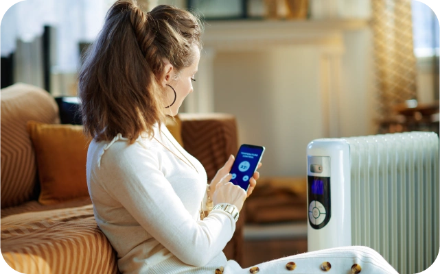 Sieviete sēž uz grīdas atspiedusies pret dīvānu ar mobilo telefonu rokās vada blakus esošā elektriskā sildītāja temperatūras iestatījumus.