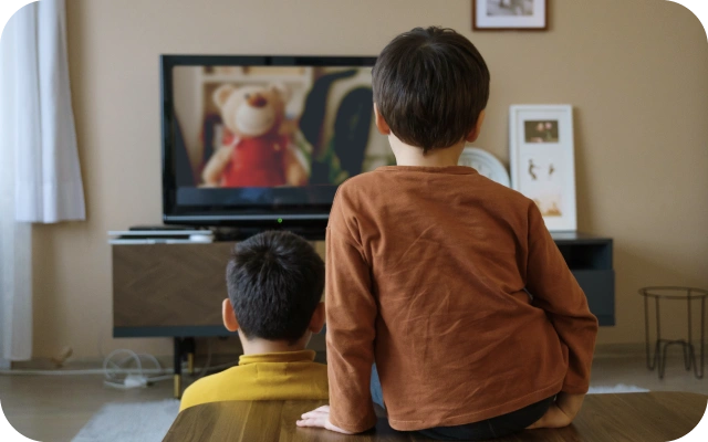 Divi bērni viesistabā skatās televizoru, kurā rāda rotaļu lāci sarkanā apģērbā; viens bērns dzeltenā džemperī sēž uz grīdas, otrs bērns brūnā džemperī sēž uz kafijas galdiņa.