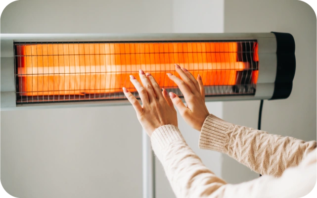 Tuvplāns sievietes rokām, kas ģērbusies gaišā džemperī, sasildās pie infrasarkanā sildītāja; fons gaiša siena.