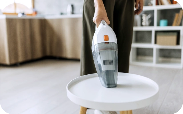 Persona tumšās biksēs izmanto baltu rokas putekļu sūcēju, lai notīrītu apaļu, baltu galdu — ierīcei caurspīdīgs putekļu konteiners un oranža poga; fonā virtuves lete un plaukti.