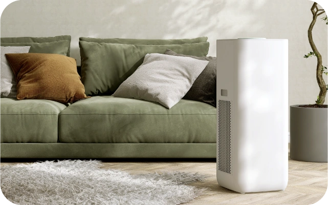 Dzīvojamā istaba ar zaļu dīvānu un spilveniem brūnos un baltos toņos; priekšplānā balts gaisa sausinātājs, fonā gaiša koka grīda un pūkains, balts paklājs.