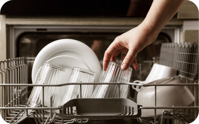 Atvērta trauku mazgājamā mašīna ar šķīvjiem, krūzēm, glāzēm un bļodām iekšpusē; roka stiepjas trauku mazgājamā mašīnā un tur vienu no glāzēm.