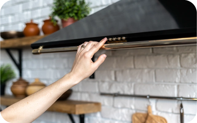 Tuvplāns ar roku, kas nospiež vienu no 5 pogām uz melna tvaika nosūcēja paneļa; ierīcei ir metāla apmale, fonā balta ķieģeļu siena un koka plaukti ar podiem.