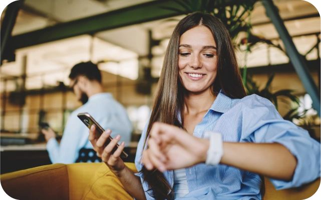 Sieviete gaiši zilā kreklā skatās uz viedpulksteni, turot otrā rokā mobilo telefonu, fonā vīrietis no mugurpuses sēž pie galda ar telefonu rokā.